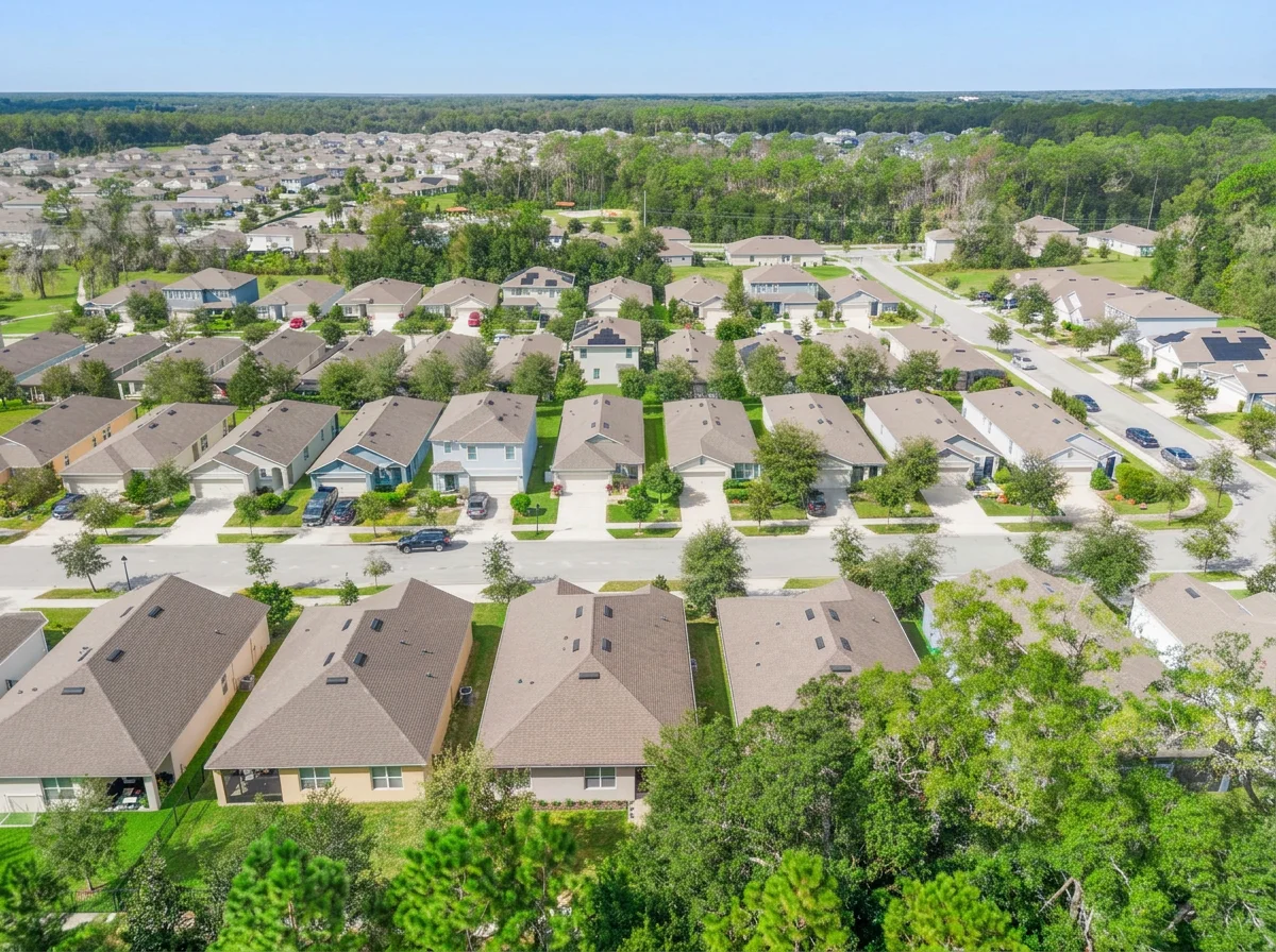 An aerial view of a suburban neighborhood with single-family homes, green lawns, trees, and parked cars on the streets.