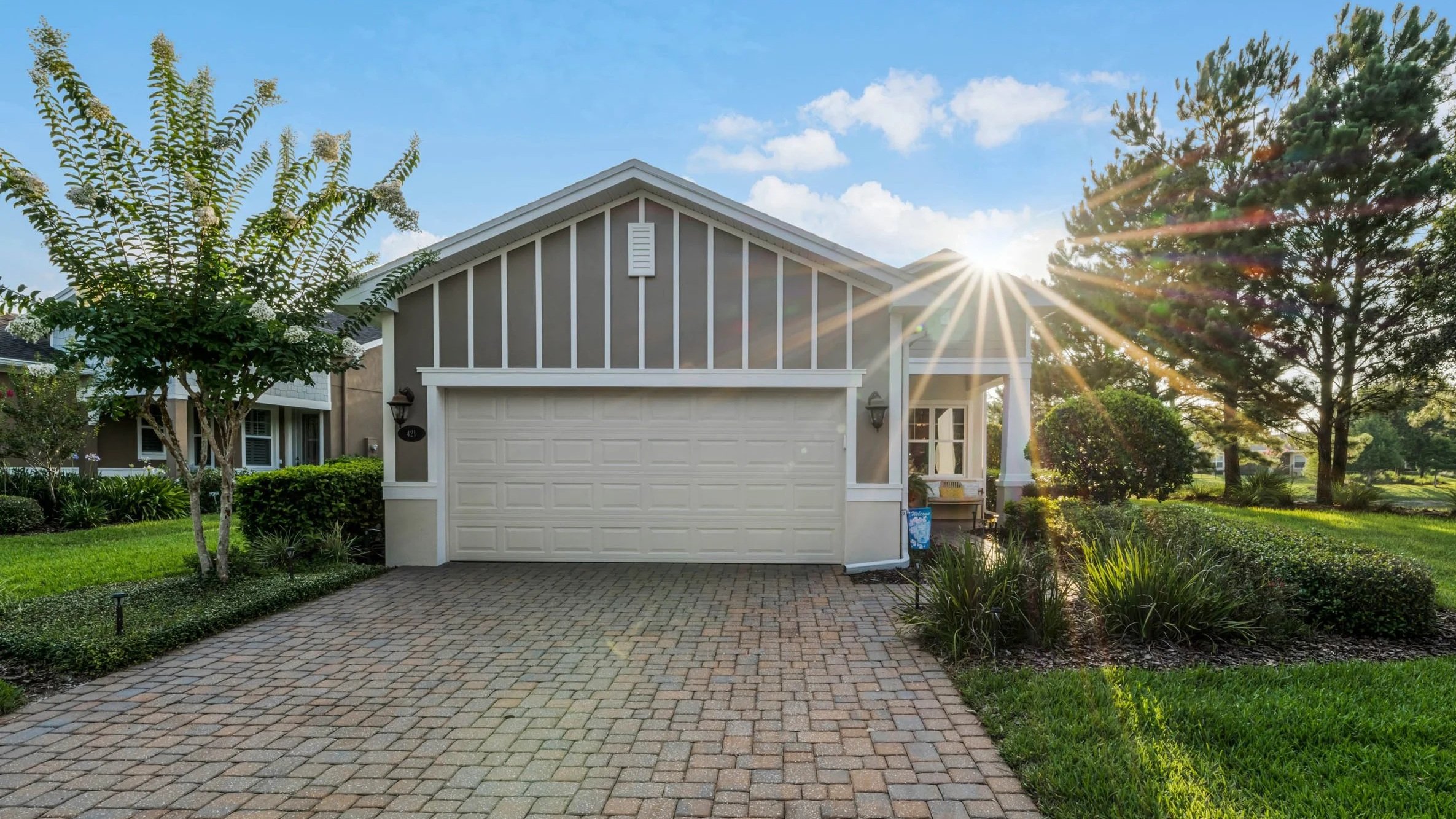 Front view of a suburban house with a white garage door, surrounded by greenery and trees, with sunlight shining through and a blue sky with some clouds.