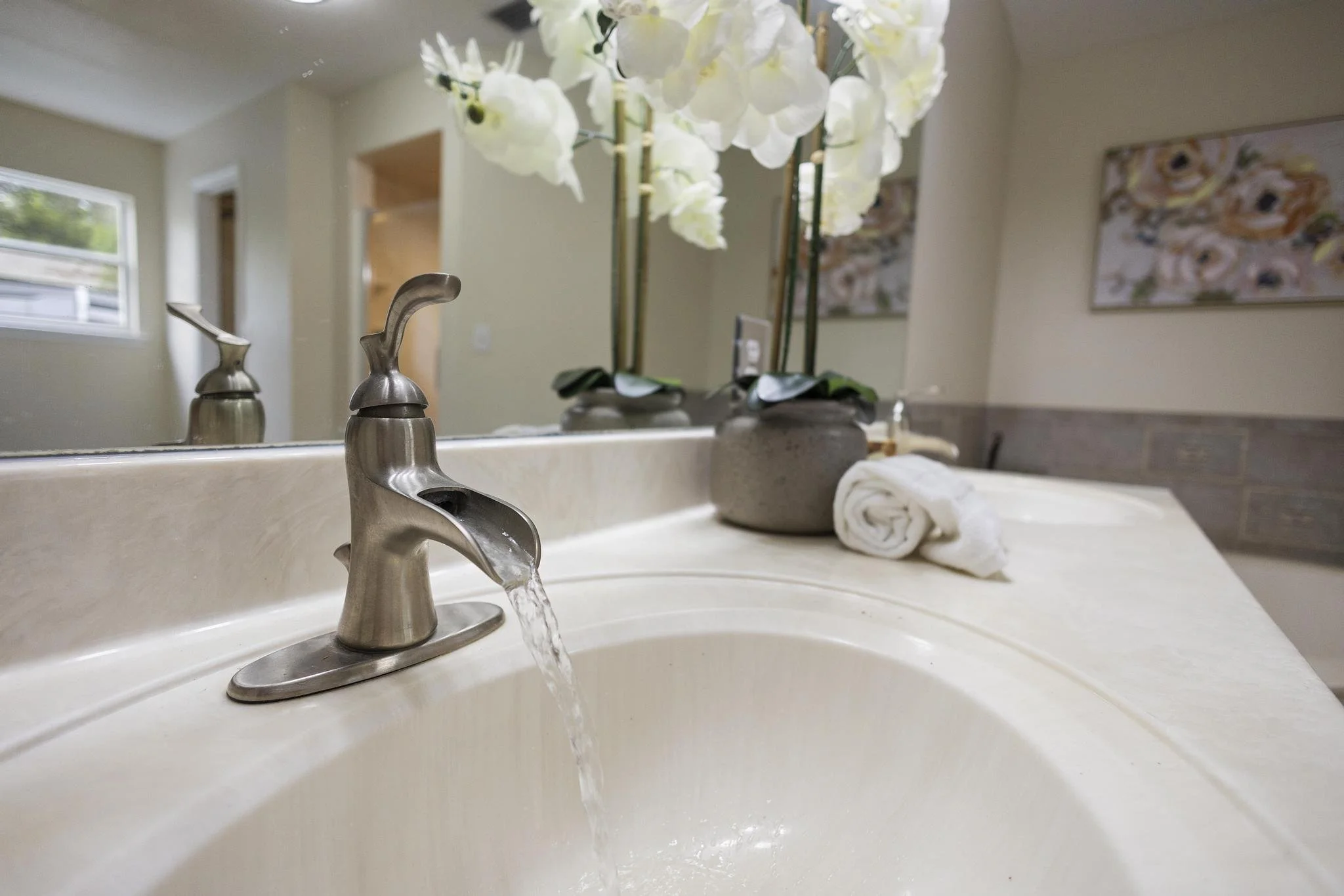 Bathroom sink with a silver faucet, a rolled white towel, and a beige flowerpot with white orchids on the countertop. In the background, a wall with a painting and window with greenery outside.
