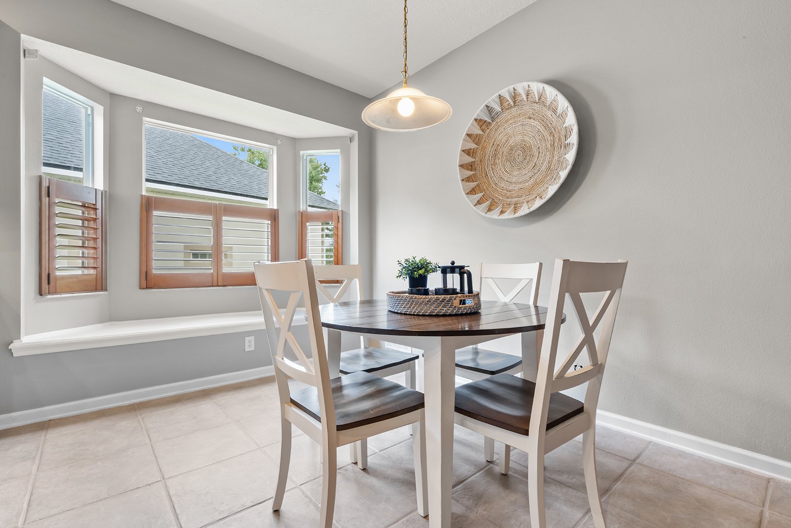 A dining area with a round wooden table surrounded by four white chairs, a tray with plants and coffee accessories on the table, a large circular wall art, and three windows with wooden shutters in a bright room.