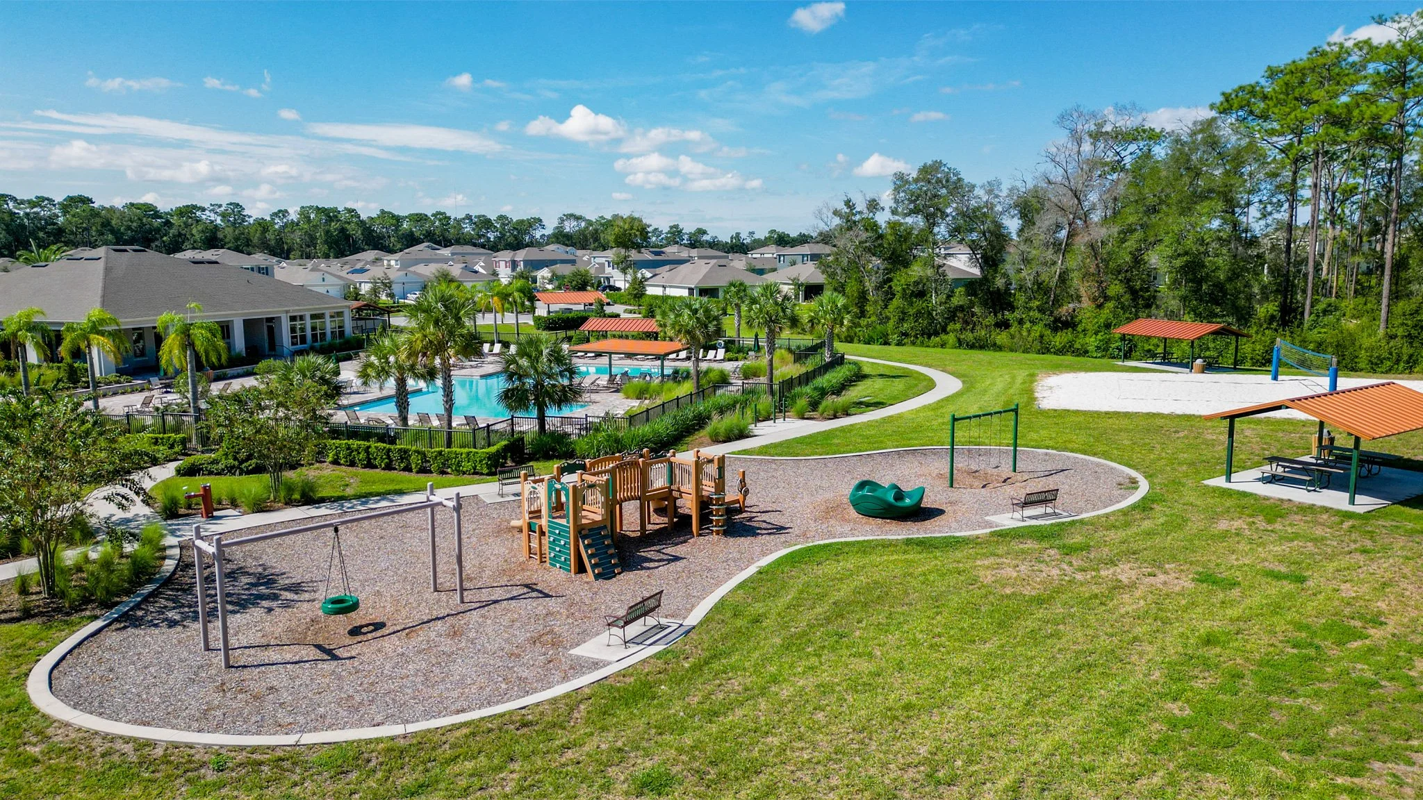 Family-friendly community park with playground, swings, picnic areas, swimming pool, surrounded by residential houses and trees, under a blue sky with clouds.
