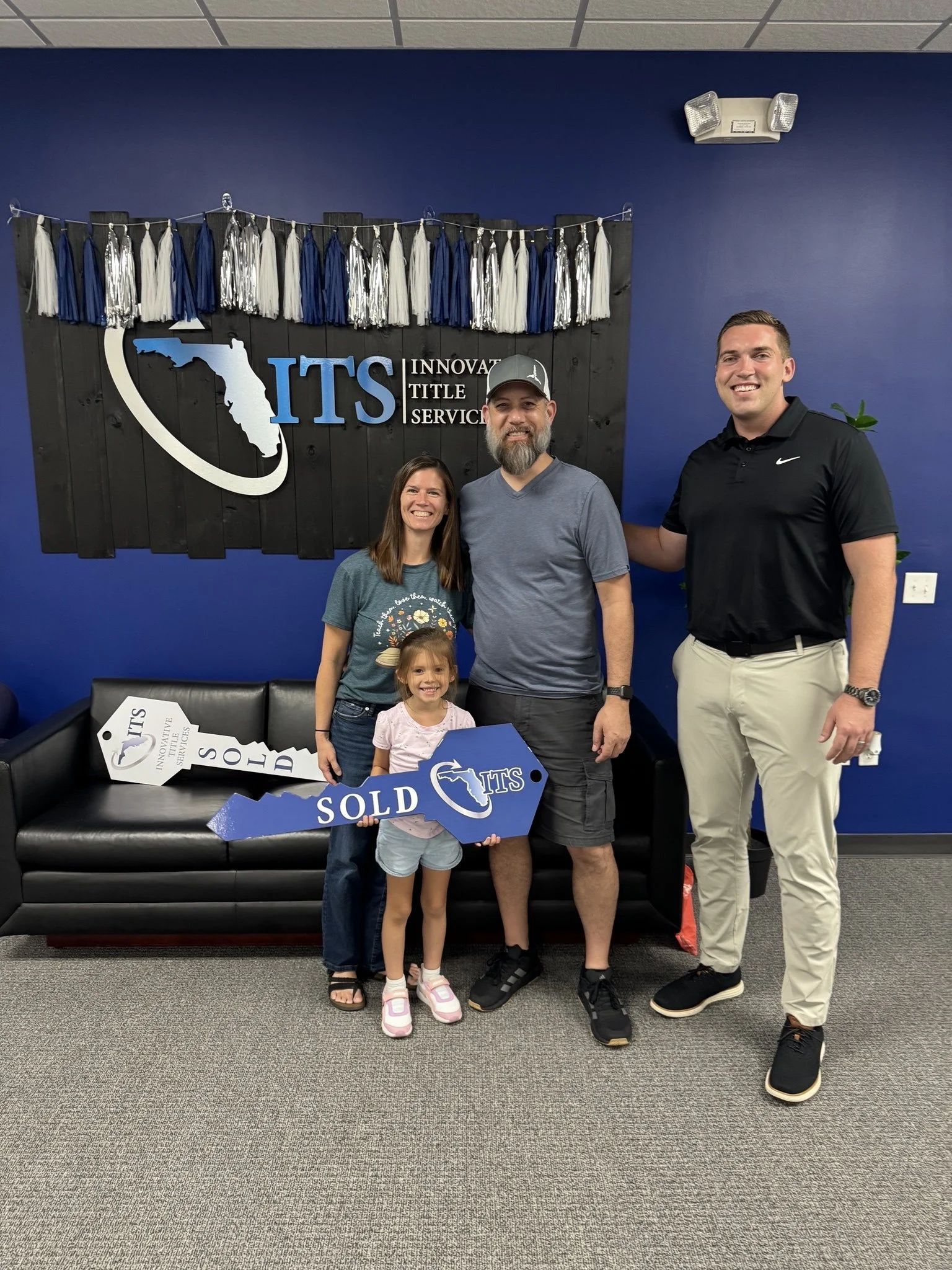 Four people, including a young girl, posing for a photo in an office or retail space with a blue wall and a decorative black wooden panel with a Florida map, the words "ITS," and business name "Innovative Title Services." They are smiling, and some are holding signs that say "Sold" and "ITS." The background also has a fringe decoration with blue, white, and silver tassels.