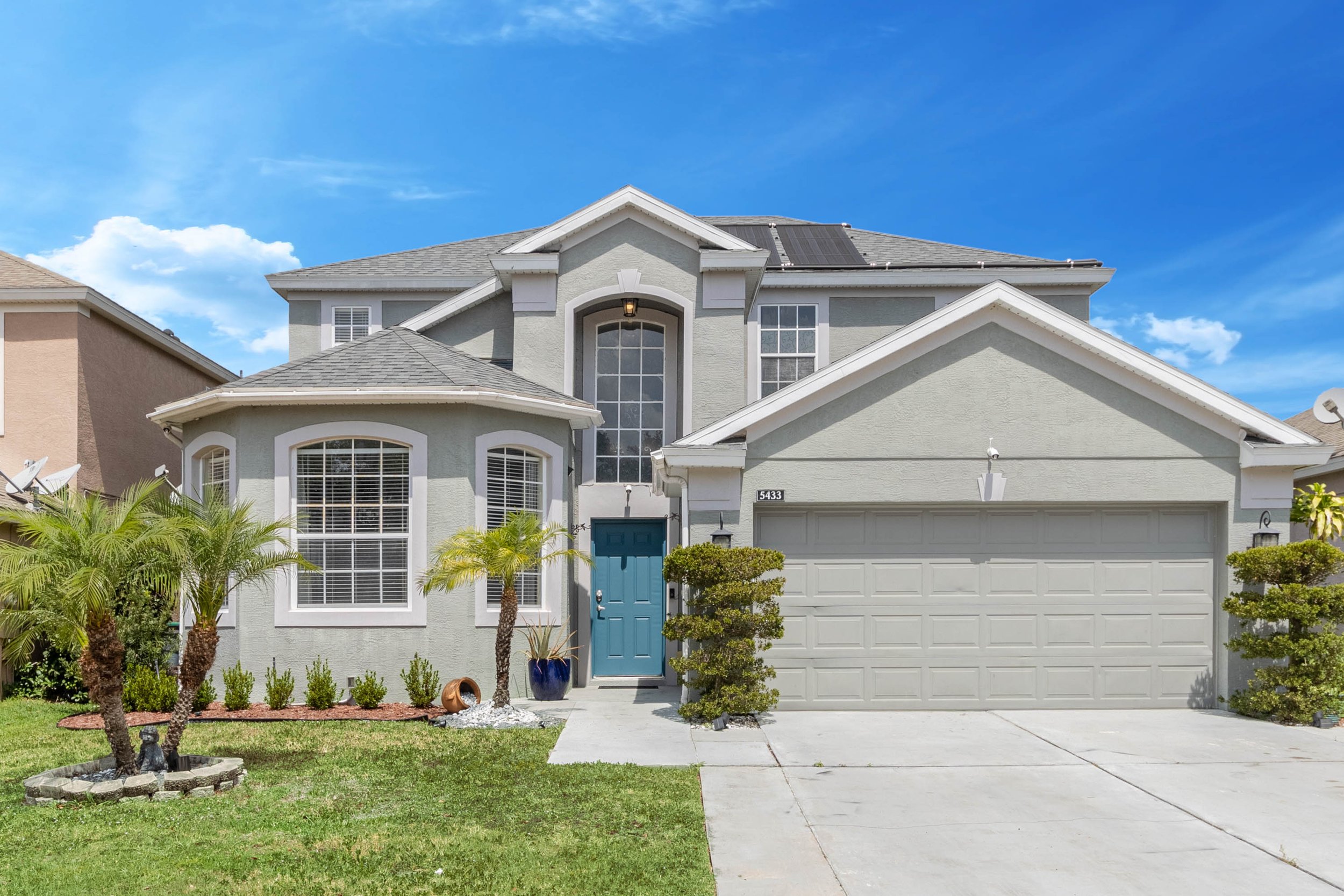 A two-story house with a gray exterior, a blue front door, large windows, a white garage door, and solar panels on the roof. Front yard with green grass, small trees, and plants under a blue sky.