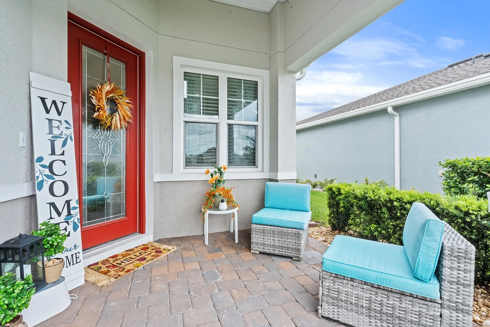 Front porch with a red door, wicker chairs with blue cushions, a small table with flowers, greenery, and a welcome sign.