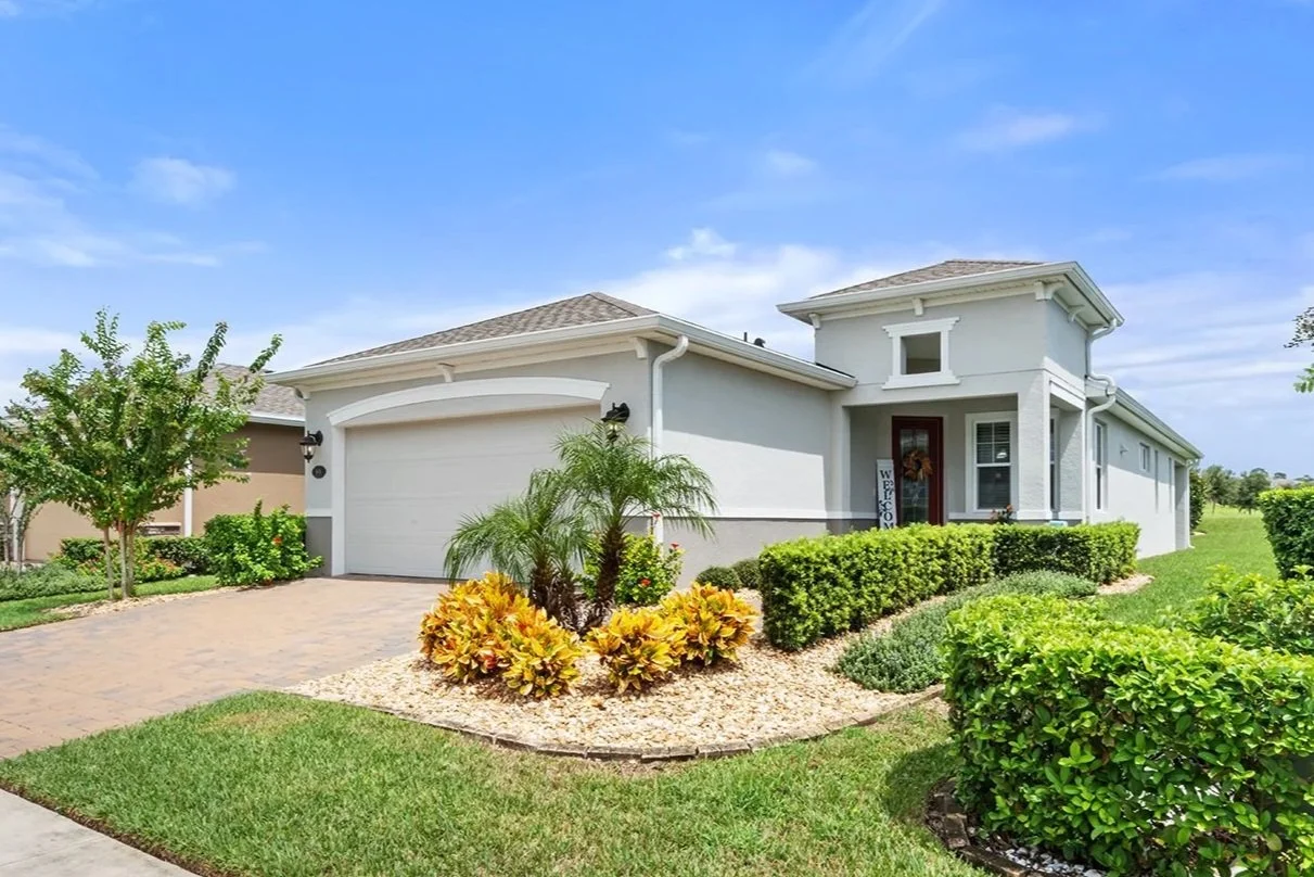 Exterior of a modern house with a two-car garage, landscaped lawn, palm trees, and shrubs under a blue sky.