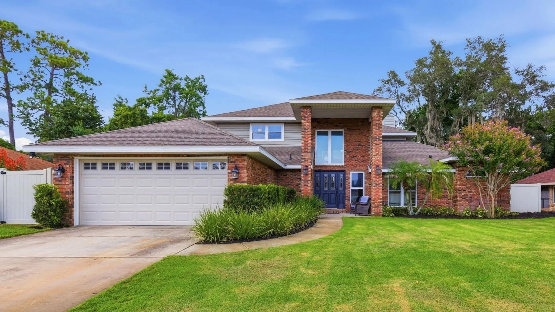 A two-story brick house with a gray shingle roof, white garage door, and a landscaped front yard with green grass, shrubs, and trees, under a blue sky with some clouds.