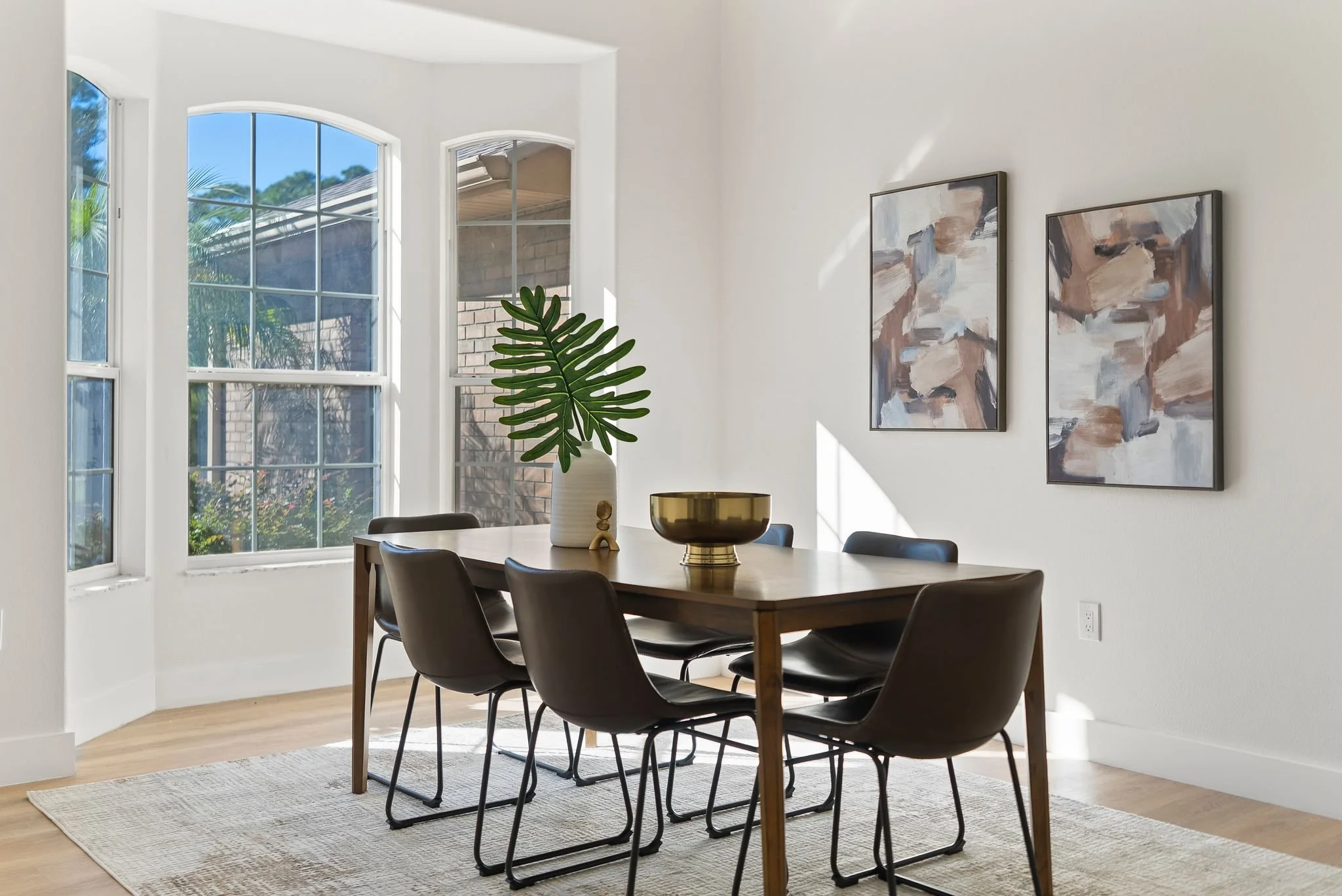 Modern dining area with a wooden table, six black chairs, large windows natural light, a potted plant, and abstract wall art.