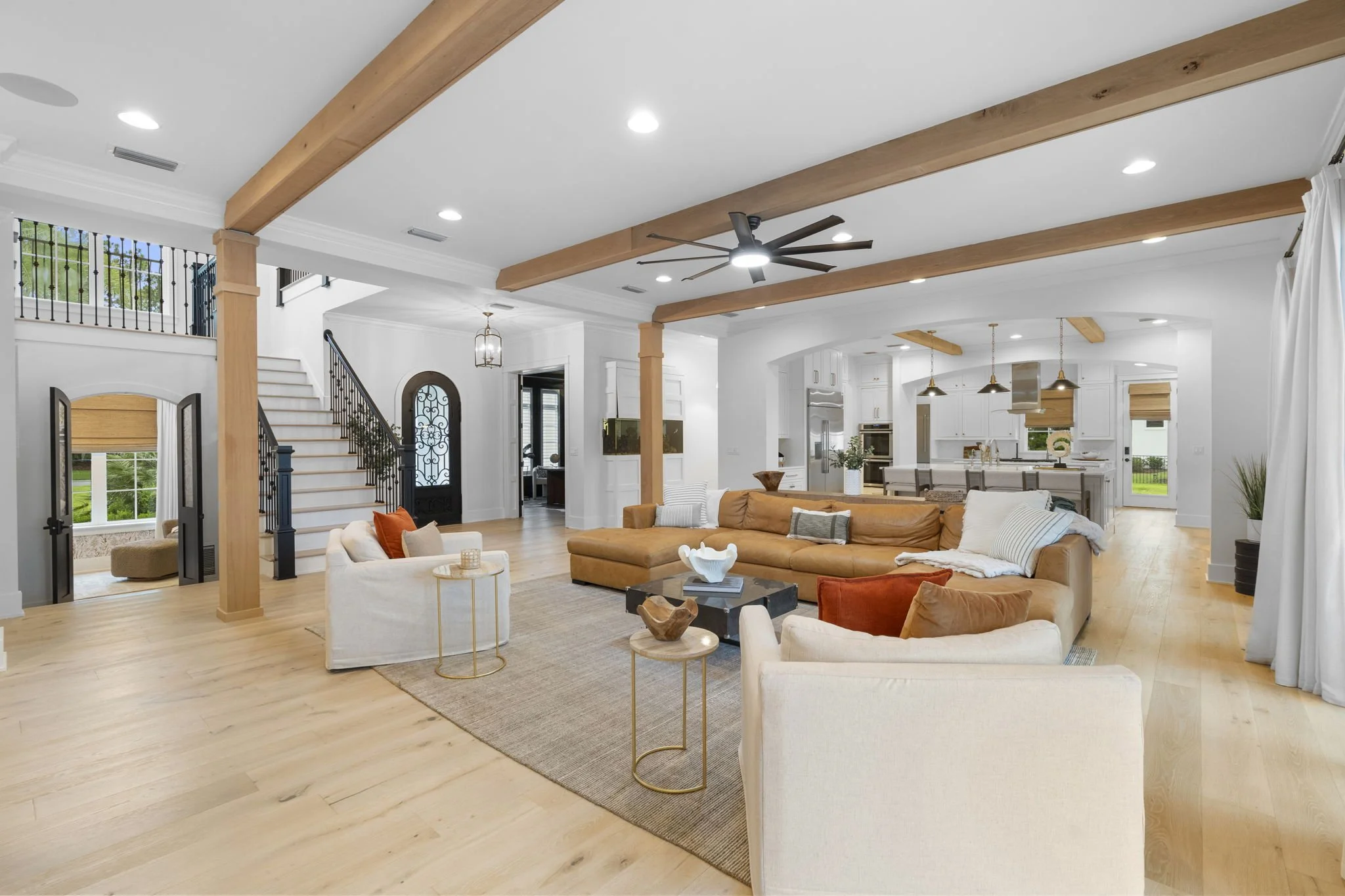 Open-concept living room and kitchen with white walls, wooden beams, a black ceiling fan, and hardwood floors. There are beige and white sofas, decorative pillows, and coffee tables. The kitchen features white cabinets, a large island, and pendant lights. Windows and a staircase are visible in the background.