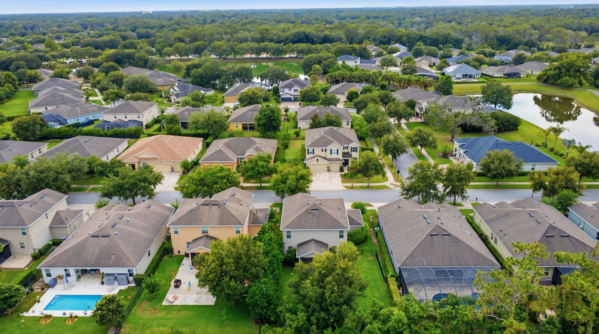 Aerial view of a suburban neighborhood with houses, trees, and greenery, some with swimming pools in backyards, and a pond surrounded by trees.