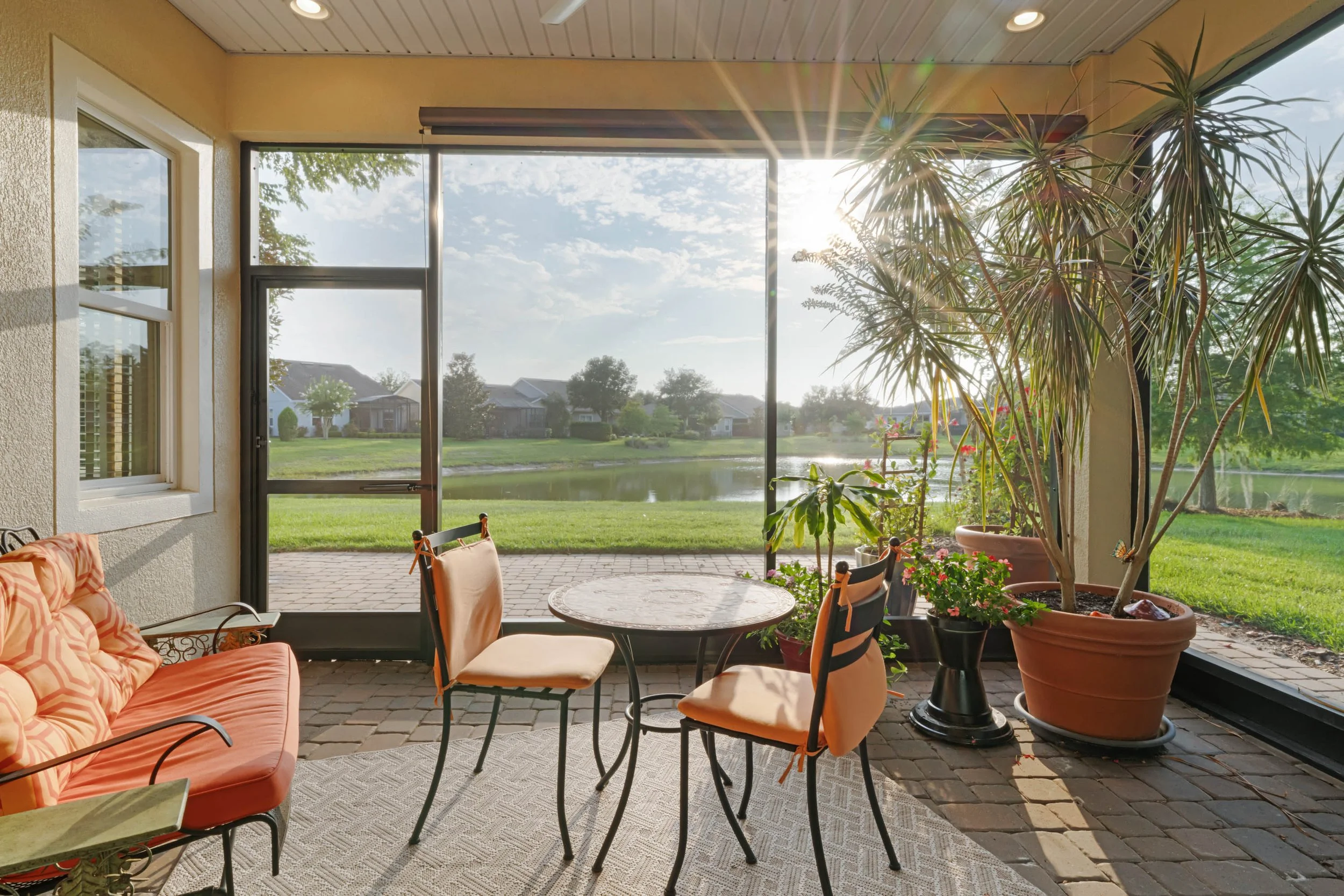 Patio area with round table and chairs, large potted plants, and a view of a pond and houses through a screened-in porch, with sunlight streaming in.