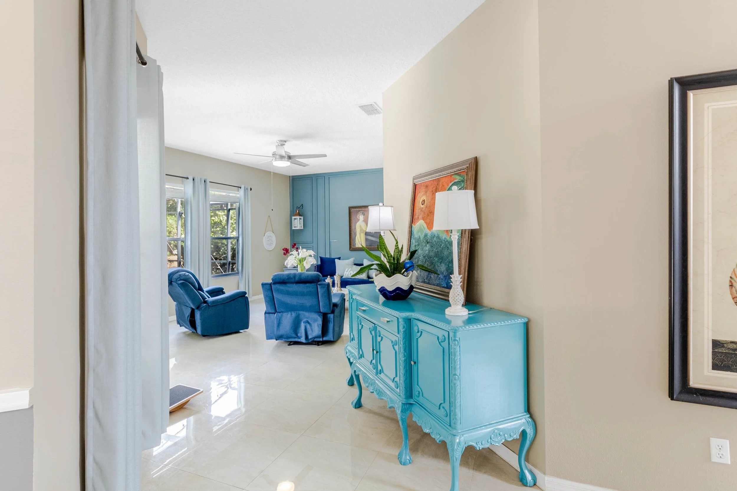 Living room with blue chairs and a teal sideboard, decorated with plants, lamps, and framed artwork, with large windows and white curtains.