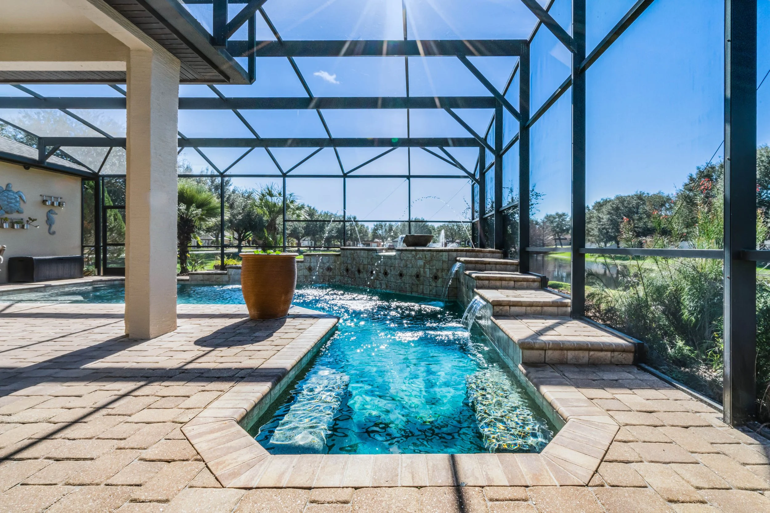 A screened-in backyard pool area with a rectangular pool featuring a small waterfall and tiered steps. There is a round, wooden decorative element beside the pool, and the area is paved with light-colored bricks. The screen enclosure allows sunlight to stream in, with trees and a bright blue sky visible outside.