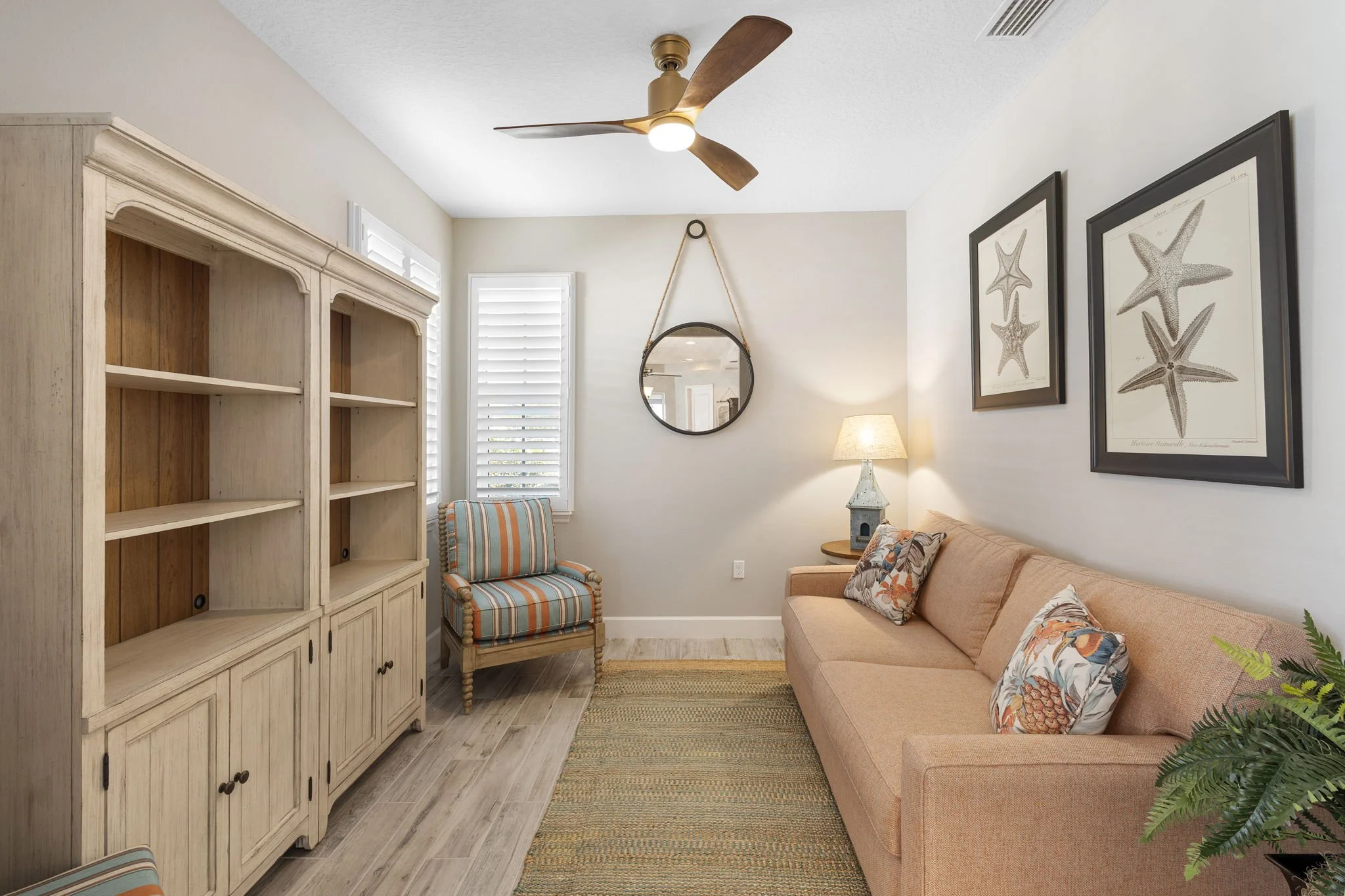 A living room featuring a beige couch with patterned pillows, a striped armchair, a wooden shelf, and wall art of starfish. There's a round mirror hanging on the wall, a table lamp, and a ceiling fan with wooden blades and a light. The room has white walls, plantation shutters on the windows, and a hardwood floor with a woven rug.