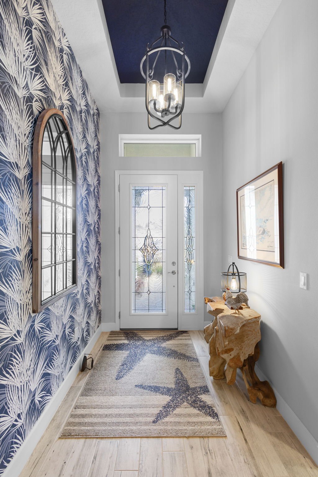 Foyer with coastal decor, patterned wall with palm leaf design, decorative wooden table with lamp and bird sculpture, starfish-patterned rug, front door with glass panels, framed artwork on the wall, modern chandelier.