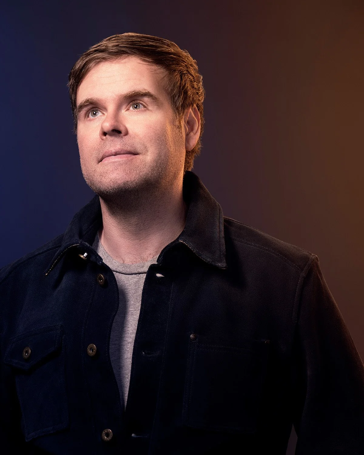 Portrait of a vocal coach with short brown hair wearing a black jacket over a gray shirt, looking upwards against a dark gradient background.