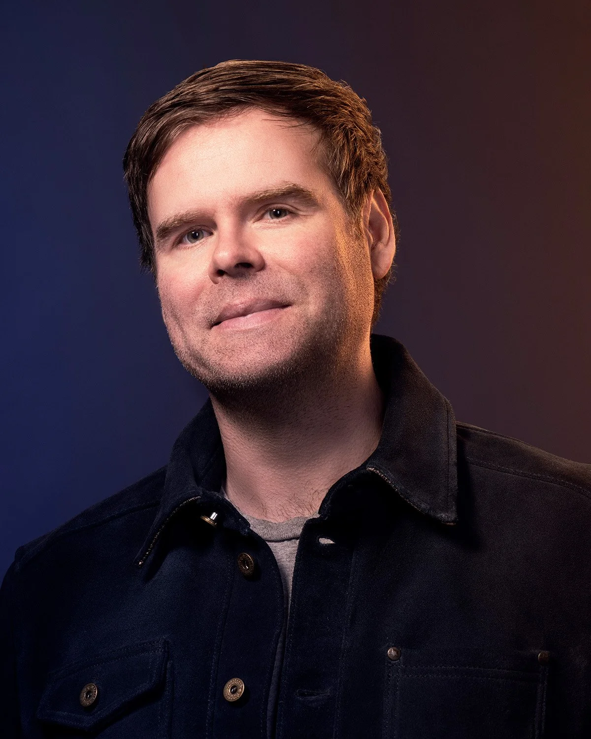 A man with short brown hair and light skin, wearing a dark button-up jacket over a light shirt, posing against a dark gradient background.