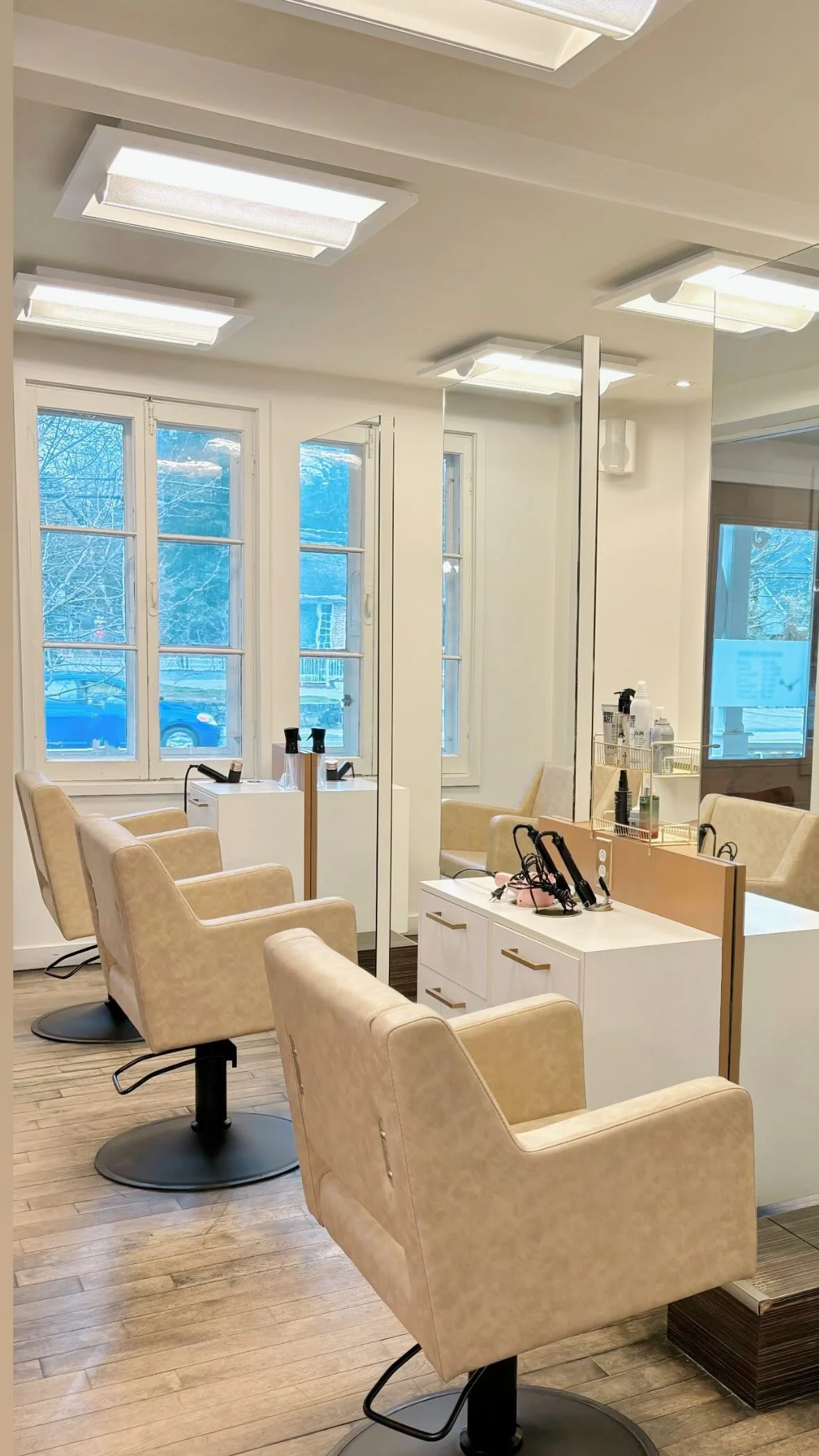 Salon de coiffure avec plusieurs fauteuils beige et miroirs, fenêtres laissant entrer la lumière naturelle, espace organisé avec outils de coiffure.