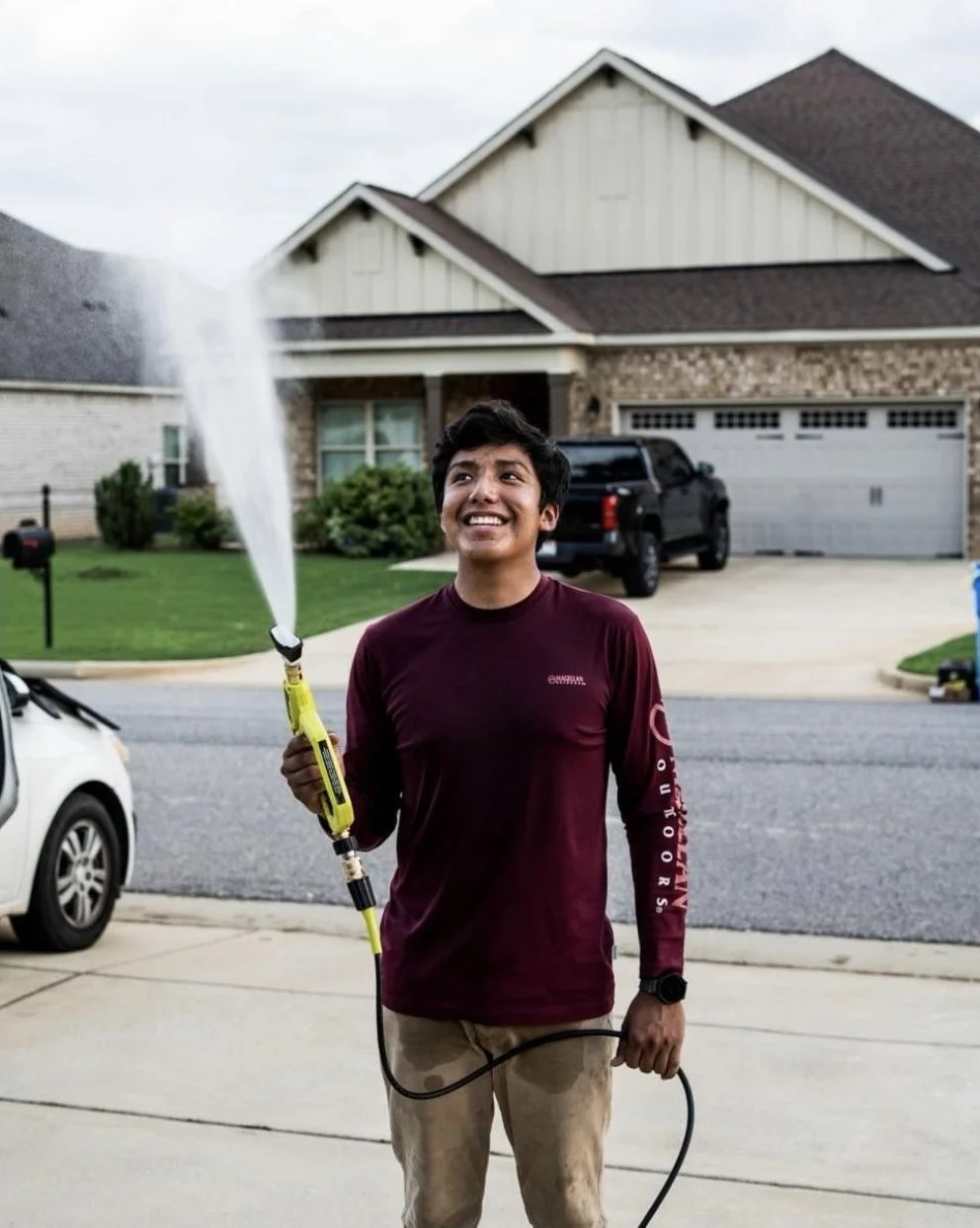 Young man watering the front yard of a suburban house with a garden hose, smiling and looking up, in a neighborhood with single-family homes and cars parked on the street.