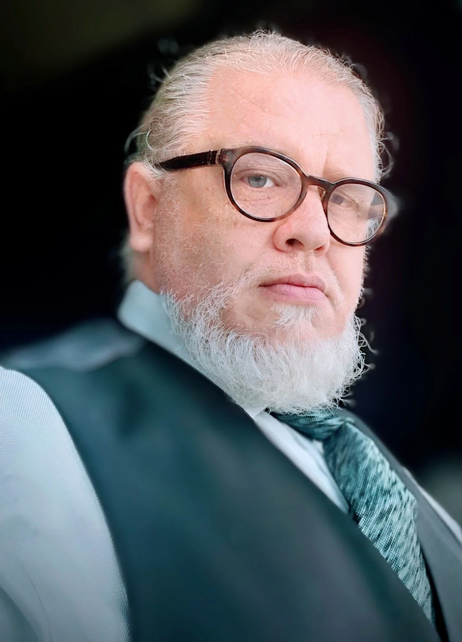 Close-up of an elderly man with gray beard and hair, wearing glasses, looking to the side with a neutral expression, dressed in a suit and tie.