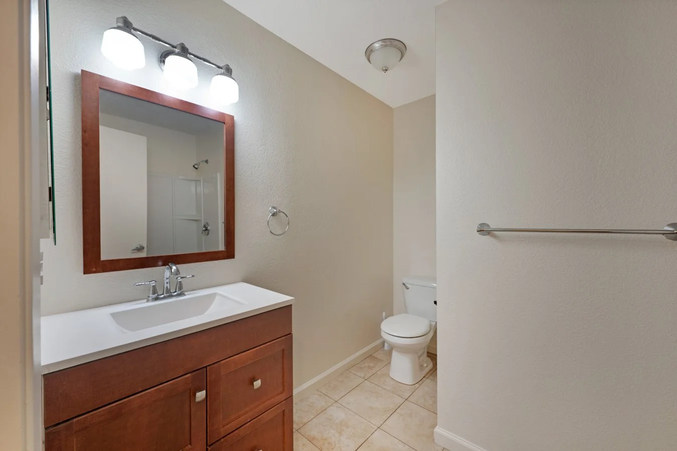 A small bathroom with a wooden vanity, mirror, and three-light fixture above it. There is a white toilet and a towel bar on the wall. Tiled floor and plain light-colored walls.