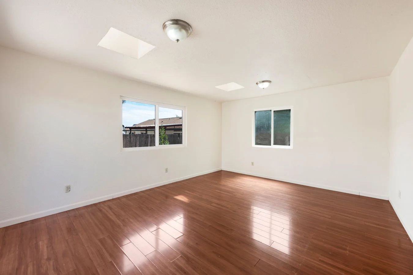 Empty living room with white walls, wood flooring, and two windows letting in natural light.