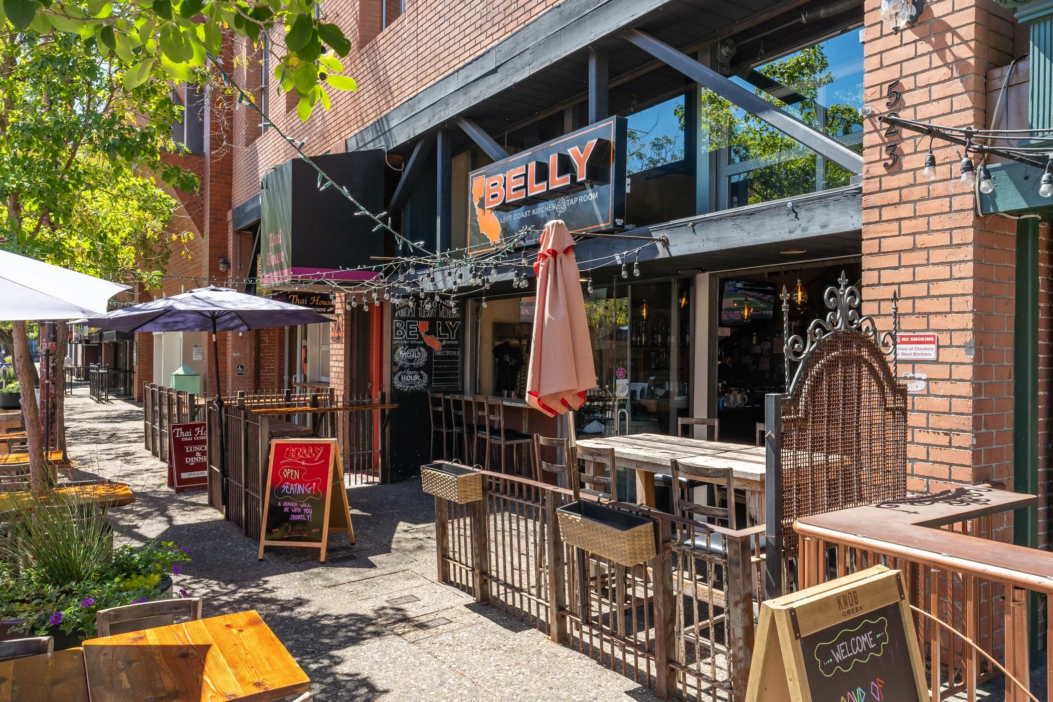 Outdoor seating area of a restaurant with wooden tables and chairs, string lights, umbrellas, and signs advertising food and seating information