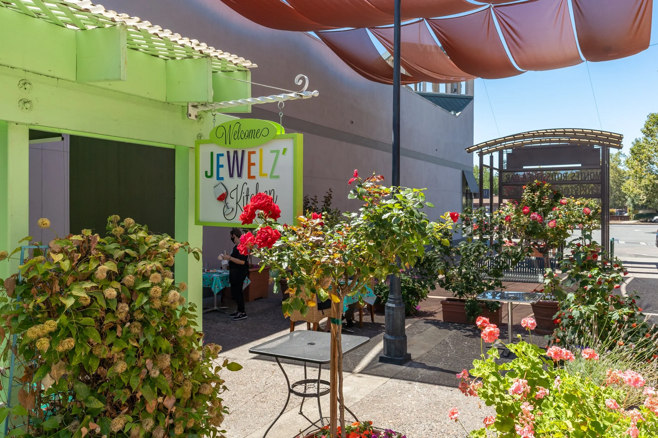 Entrance to a business called 'Jewelz' with a colorful sign, surrounded by potted plants and blooming flowers, with a partially visible outdoor setup and a person working at a table under a bright blue sky.
