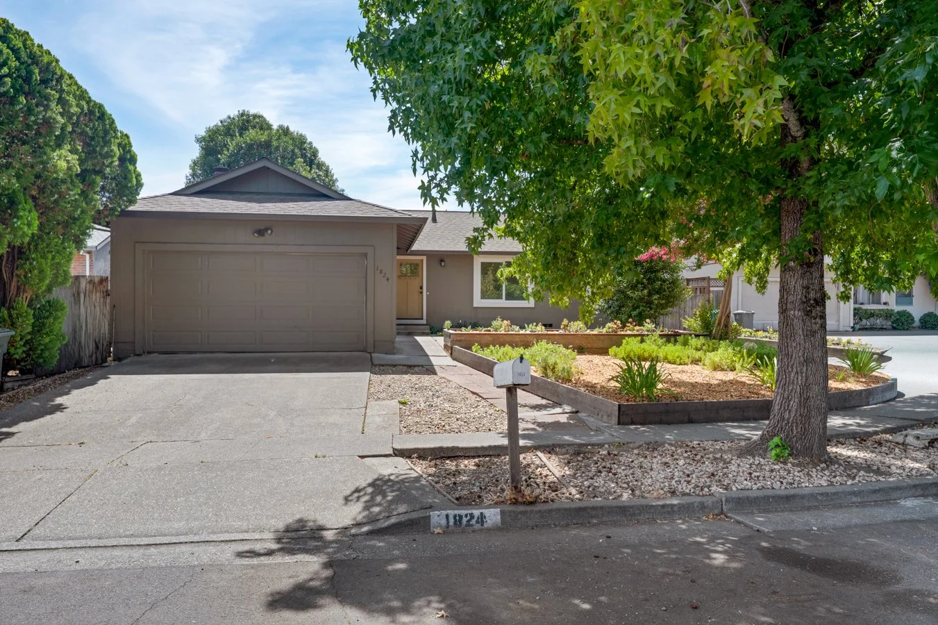 A suburban house with a gray exterior, a closed garage door, a concrete driveway, and a small front yard with a garden bed and a large leafy tree.
