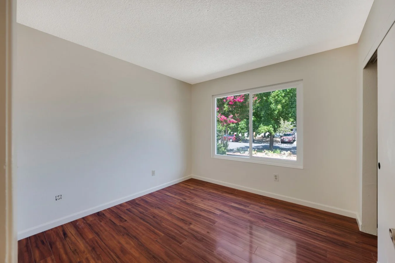 Empty room with white walls, hardwood floor, large window with views of trees and parked cars.