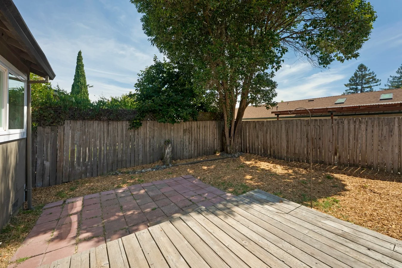 Backyard with a wooden fence, a large tree, a small paved area, and a wooden deck, under a partly cloudy sky.