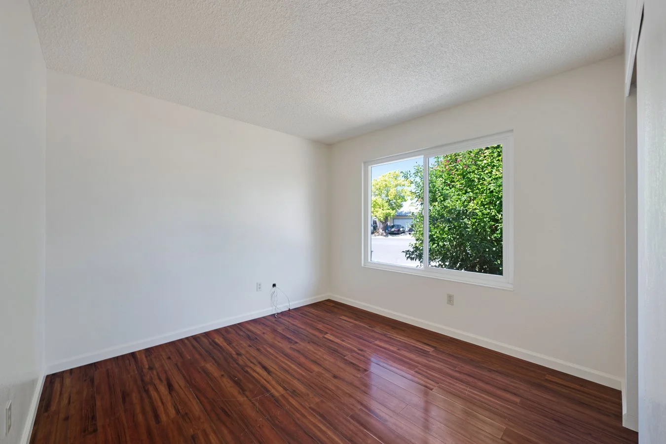 Empty room with white walls, a large window showing green trees outside, and wooden flooring.