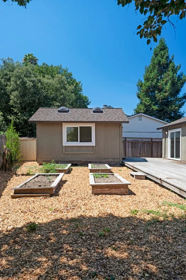 Backyard with a small beige house, raised garden beds, wooden deck, trees, and a clear blue sky.