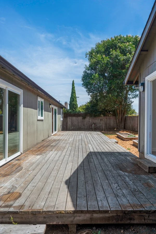 Backyard area with a wooden deck, beige house walls with sliding glass doors and windows, a tall tree, a fence, and a blue sky with some clouds.