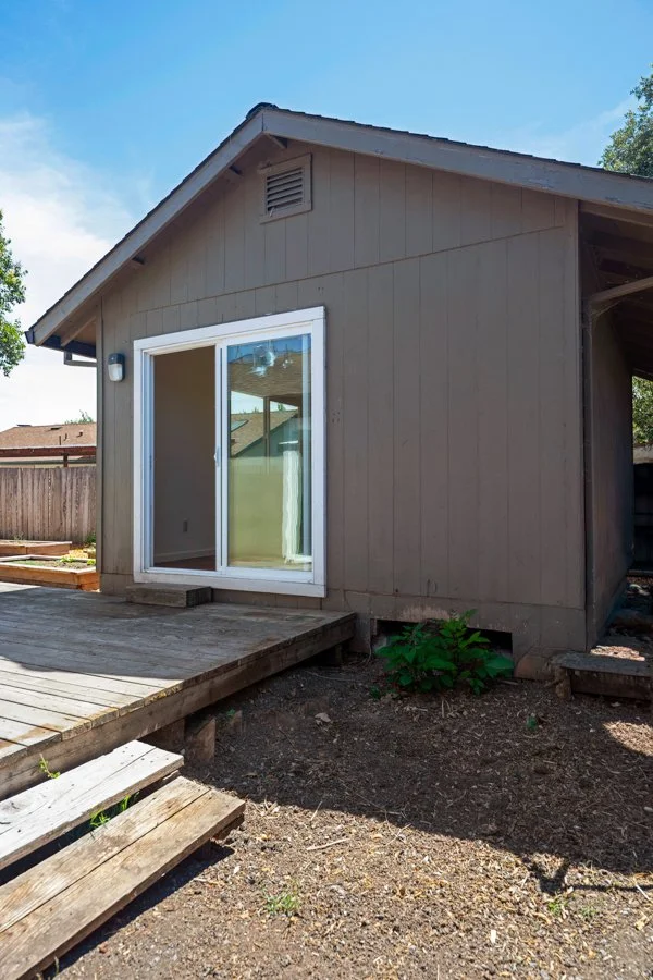 Backyard of a house with a wooden deck and sliding glass door, surrounded by a wooden fence and some plants, under a clear blue sky.