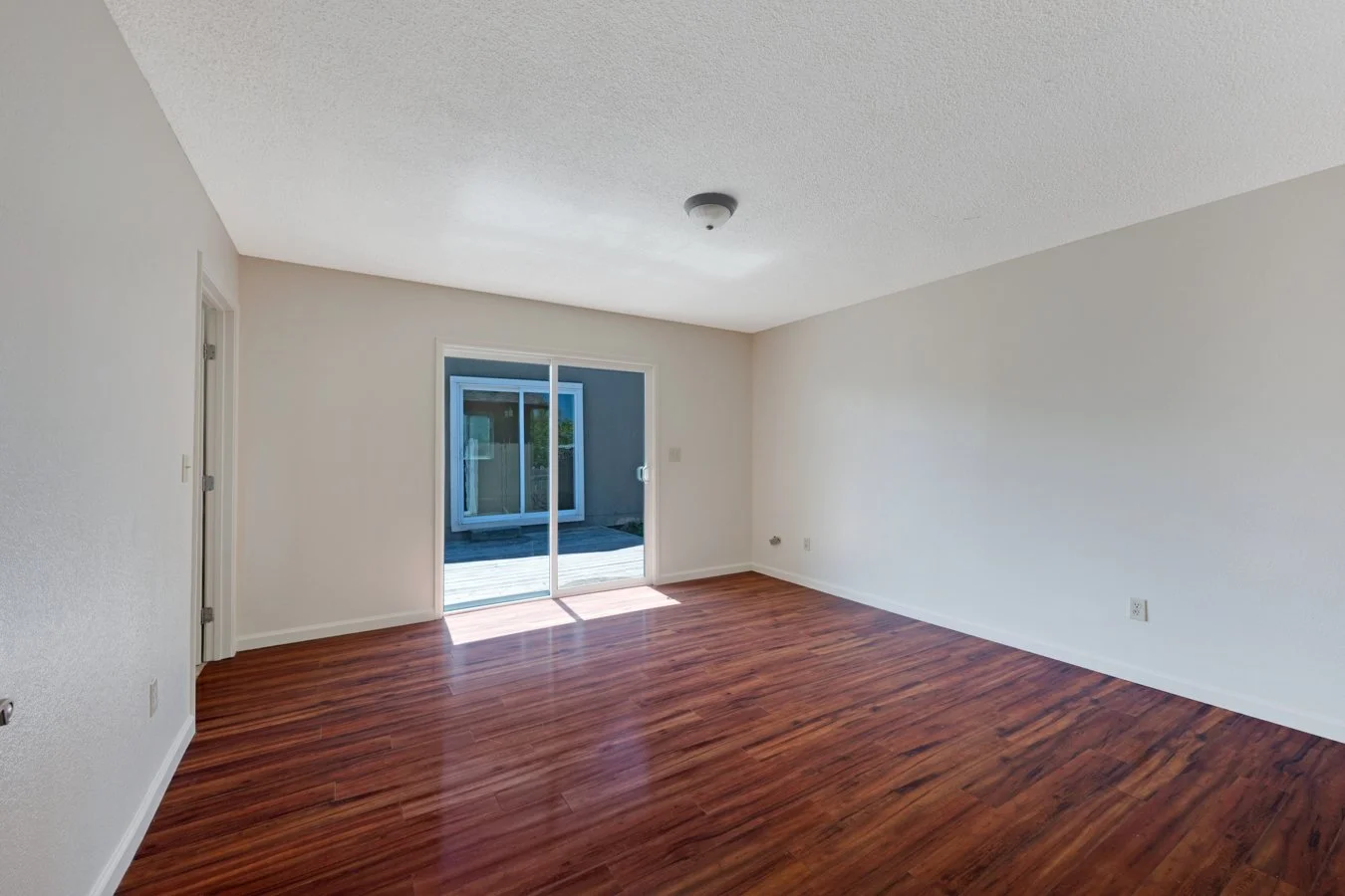 Empty living room with white walls, hardwood floors, a sliding glass door leading outside, and a ceiling light fixture.