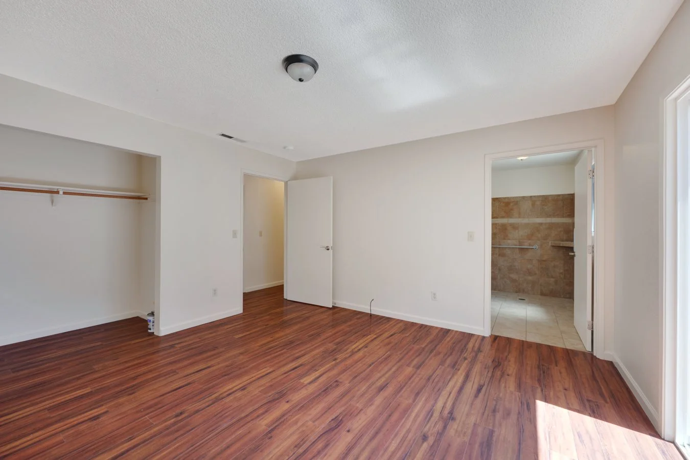 Empty room with hardwood floors, open closet, open door to bathroom with tile floor and walls, white walls, ceiling light fixture, and corner window