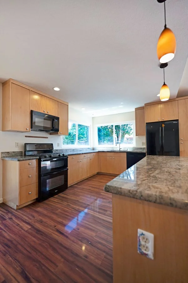Kitchen with light wood cabinets, granite countertops, hardwood floor, black appliances including a stove, microwave, and refrigerator, and large window with trees outside.