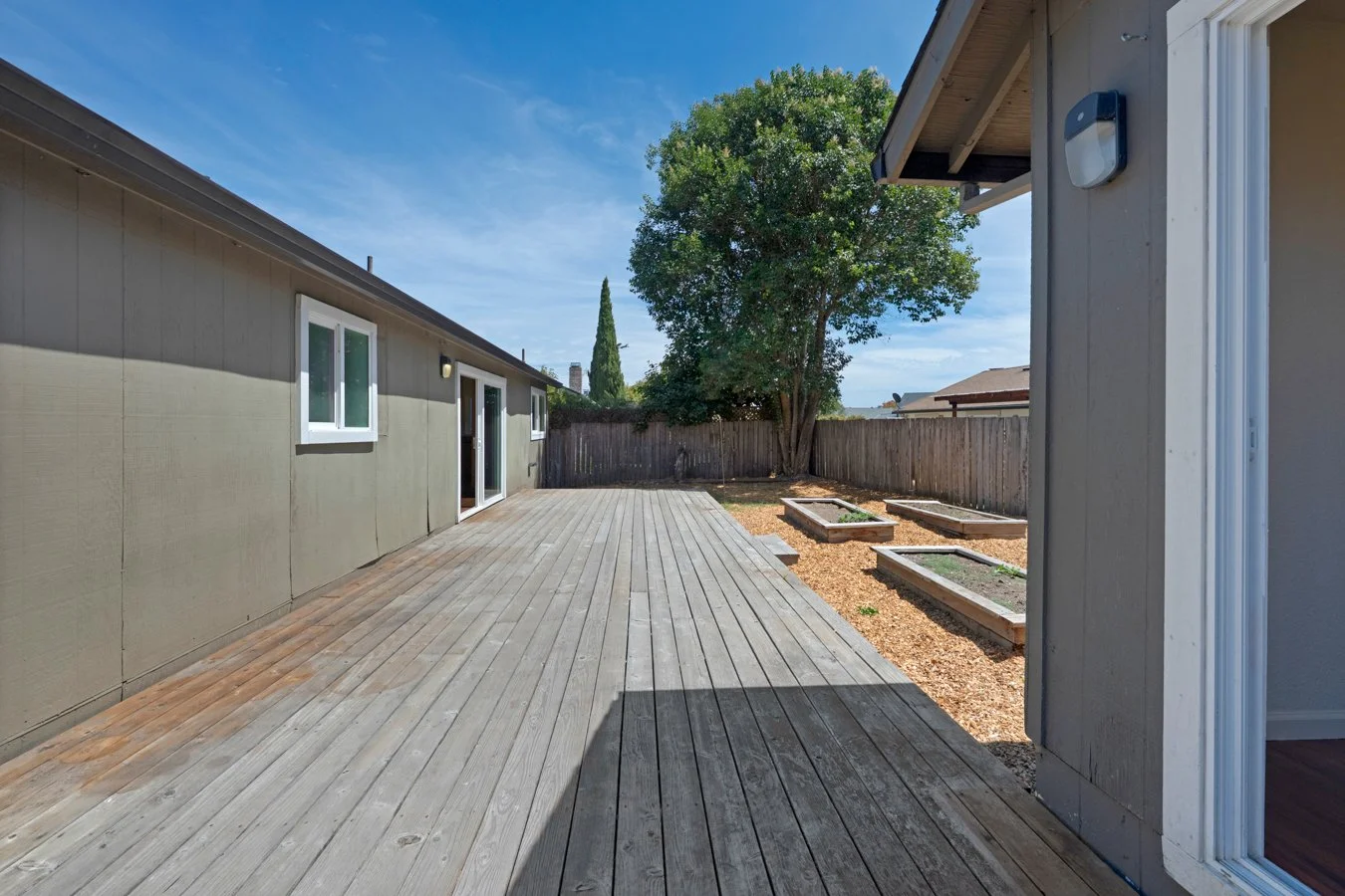 Backyard with a wooden deck, a large tree, raised garden beds, and a wooden fence under a blue sky.