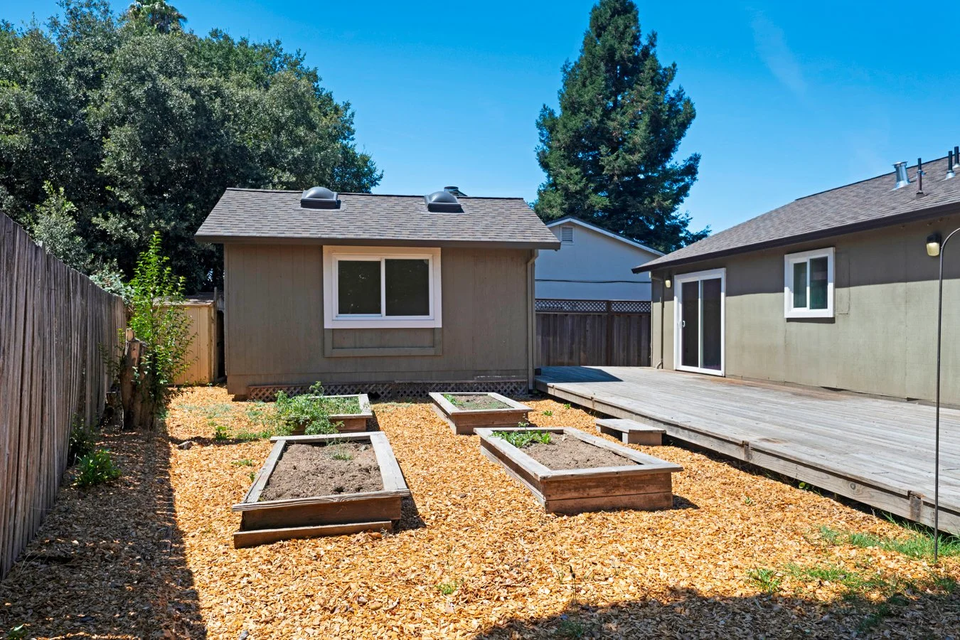 Backyard with raised garden beds, a wooden deck, and two small houses with a blue sky.