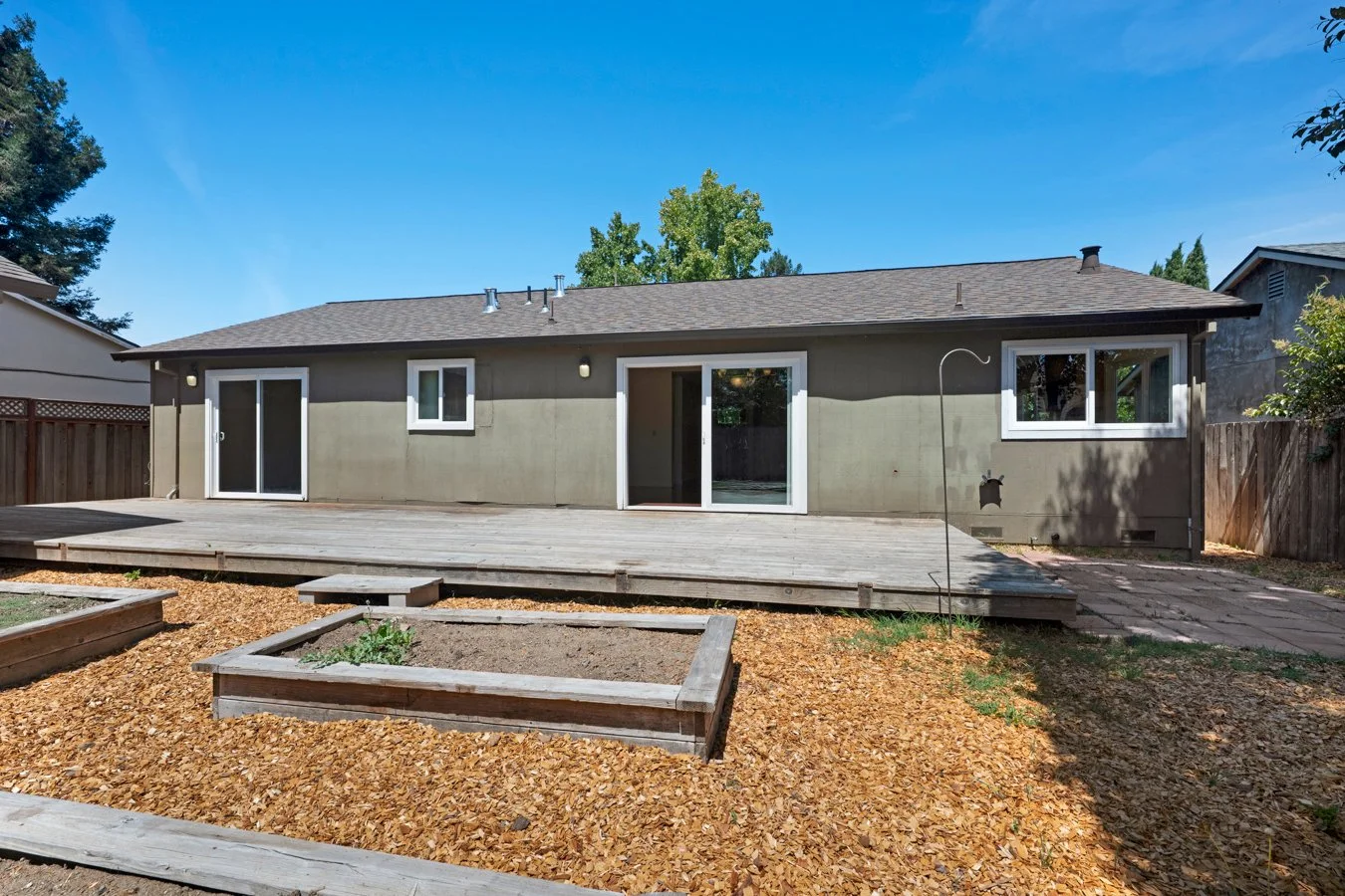Backyard with a wooden deck, garden beds, and mulch, wooden fencing, and a single-story house with sliding glass doors and windows.