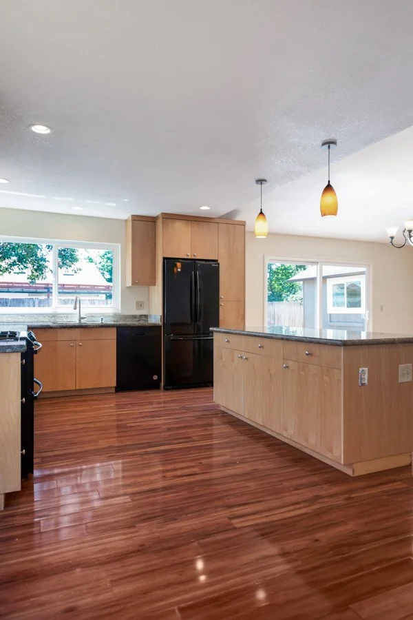 Empty modern kitchen with wooden cabinets, black appliances, granite countertops, hardwood flooring, pendant lights, and a large window.