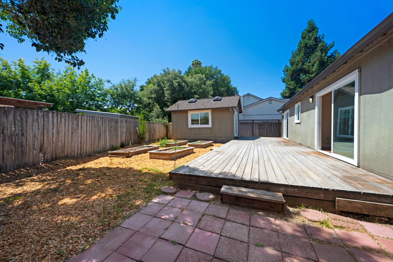 Backyard with wooden deck, paver patio, garden beds, and a wooden fence, under a clear blue sky.