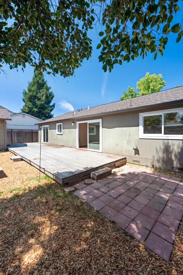 Backyard with a wooden deck, paver patio, and a beige house with sliding glass door and windows, under a clear blue sky.