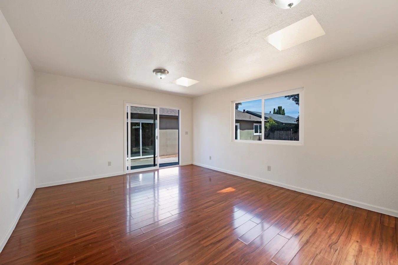 Empty living room with white walls, wooden floors, a sliding glass door leading to the backyard, and a window showing neighboring houses.