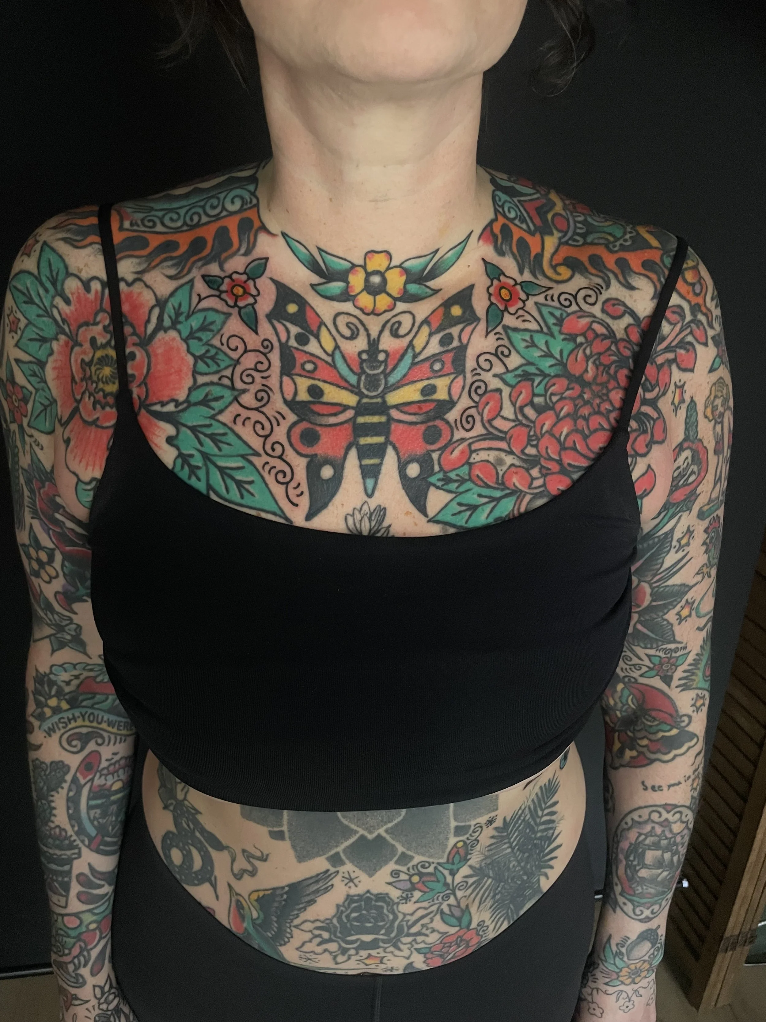 A person with extensive colorful tattoos covering their chest, arms, and stomach. The tattoos feature flowers, a butterfly, animals, and various decorative patterns.