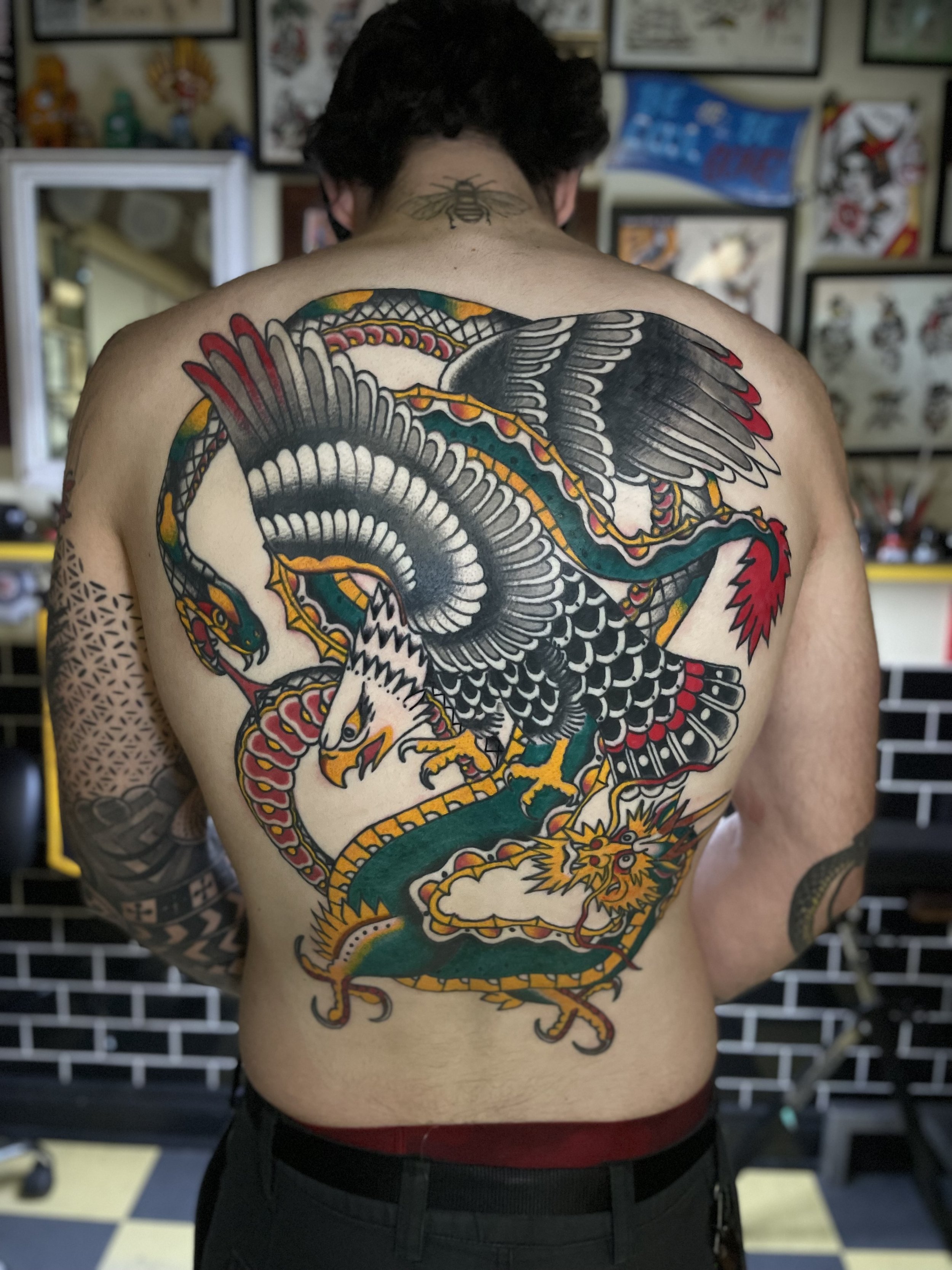 Back view of a person with a large, colorful tattoo of a dragon on their back, with an additional small bee tattoo on the neck. The background features framed artwork and posters.