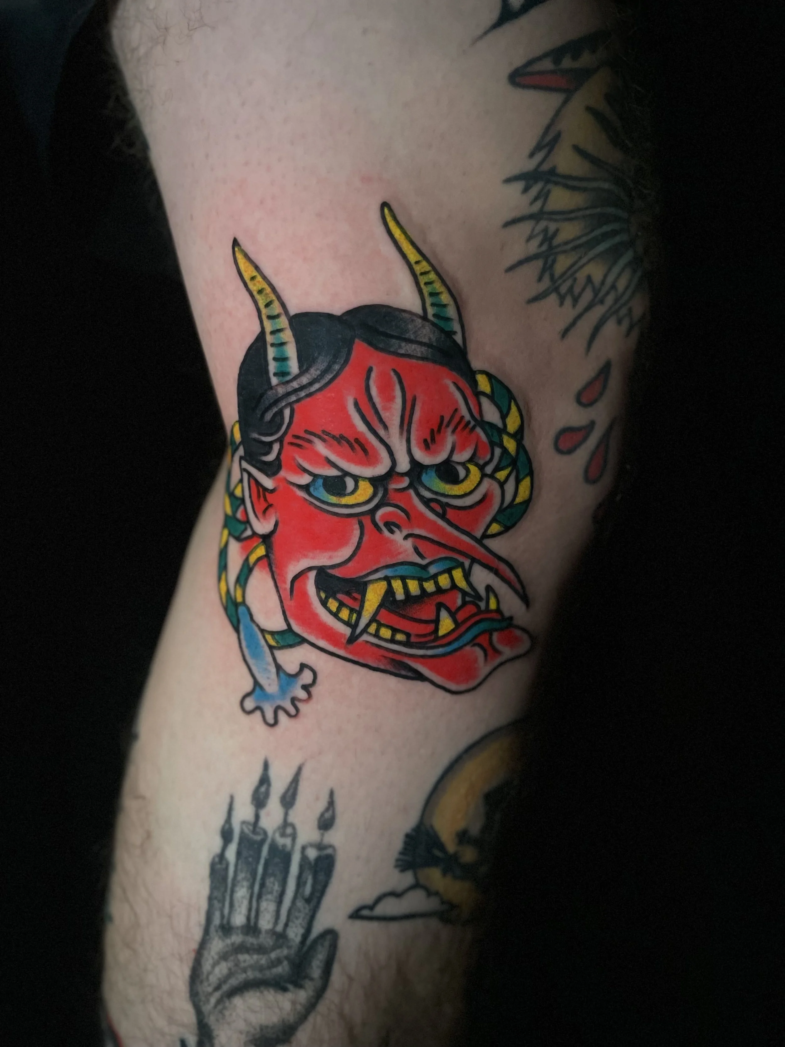 Tattoo of a traditional Japanese Hannya mask with horns, sharp teeth, and fierce eyes on a person's upper arm, with part of a dragon or snake tattoo visible above and a darker background.