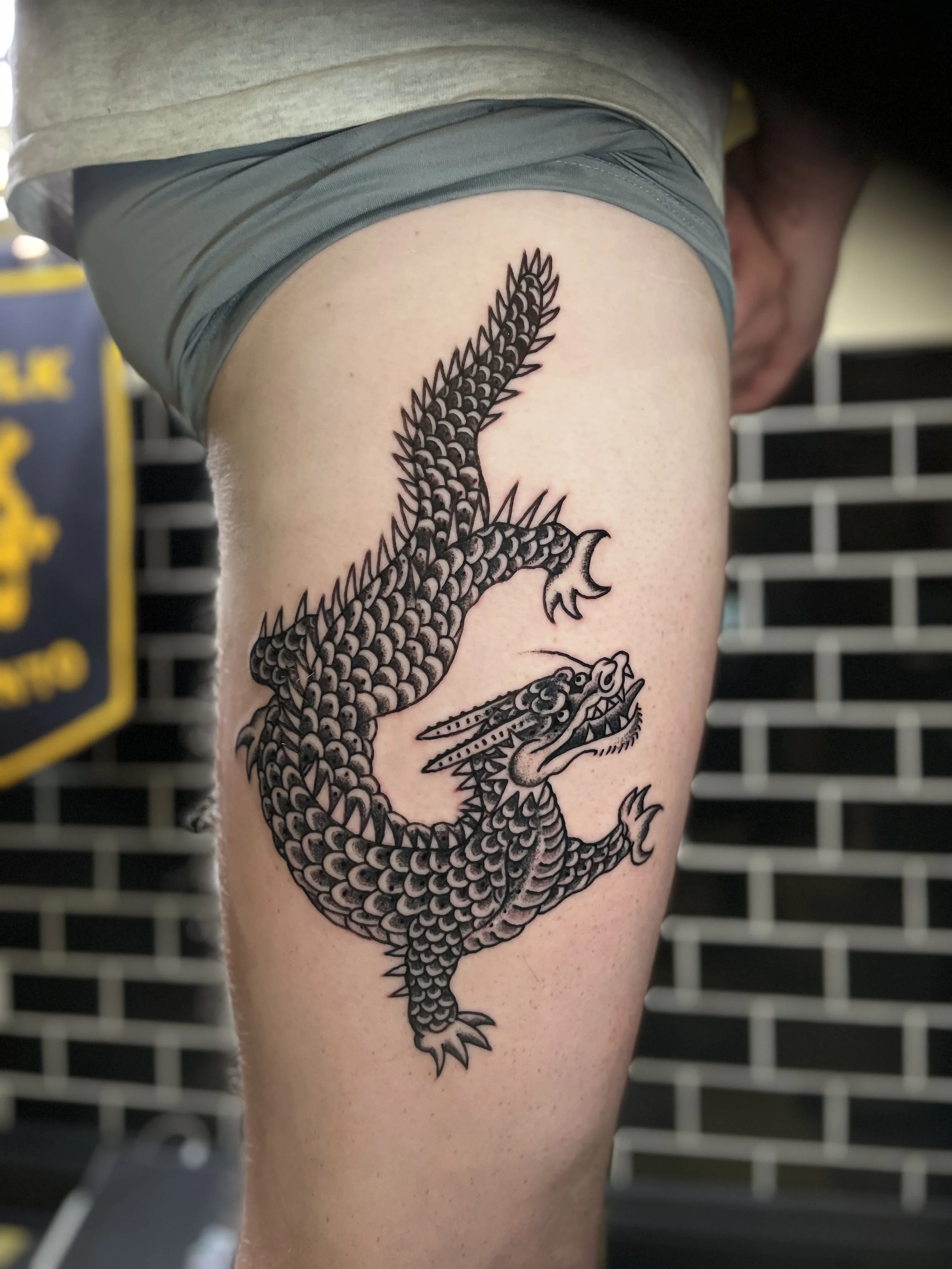 Close-up of a black ink tattoo of a dragon on a person's thigh, with details of the scales, claws, and open mouth, against a blurred background of a gym or tattoo studio.