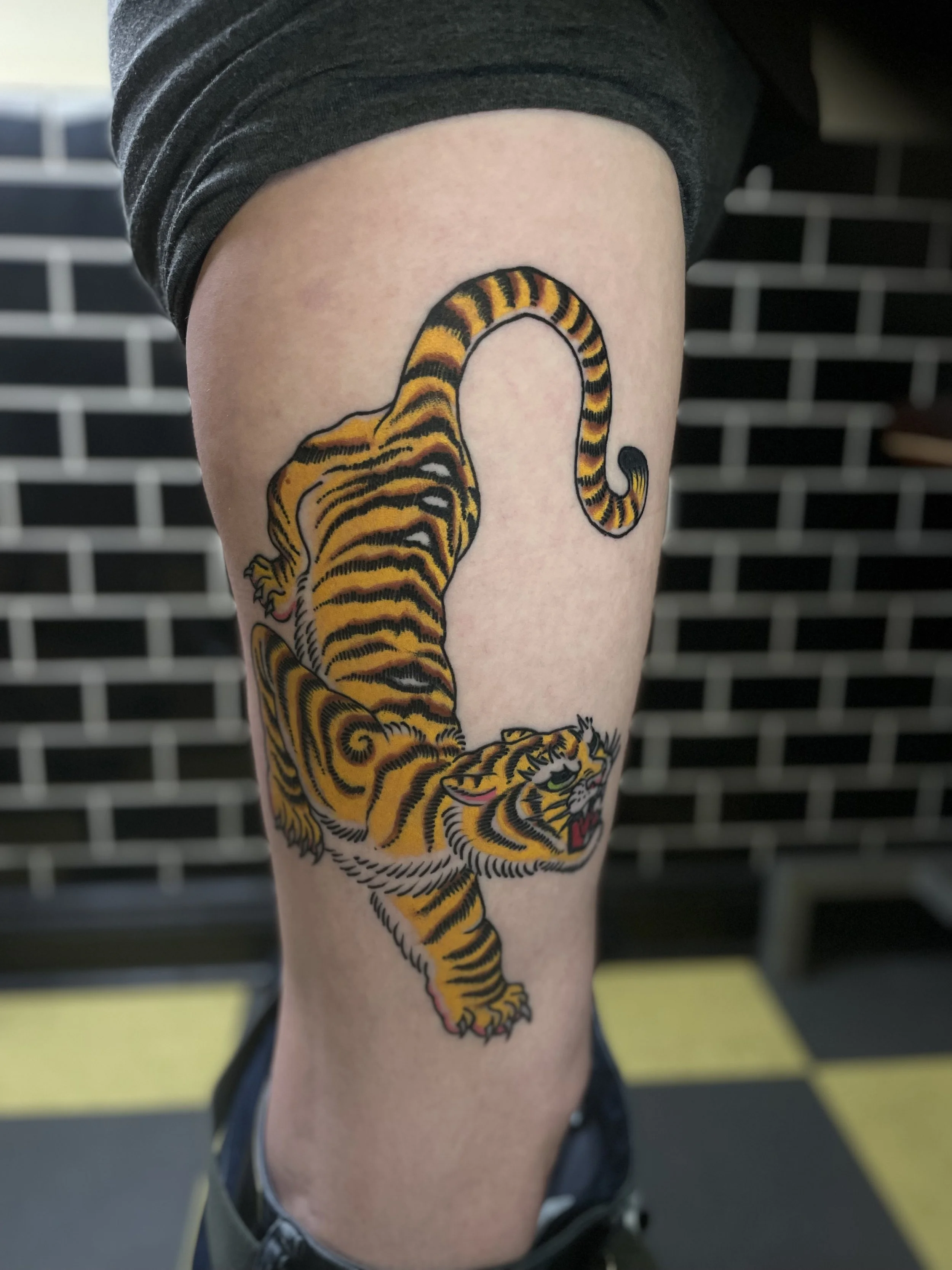 Tattoo of a yellow and black tiger on a person's thigh, with a black and white brick wall in the background.