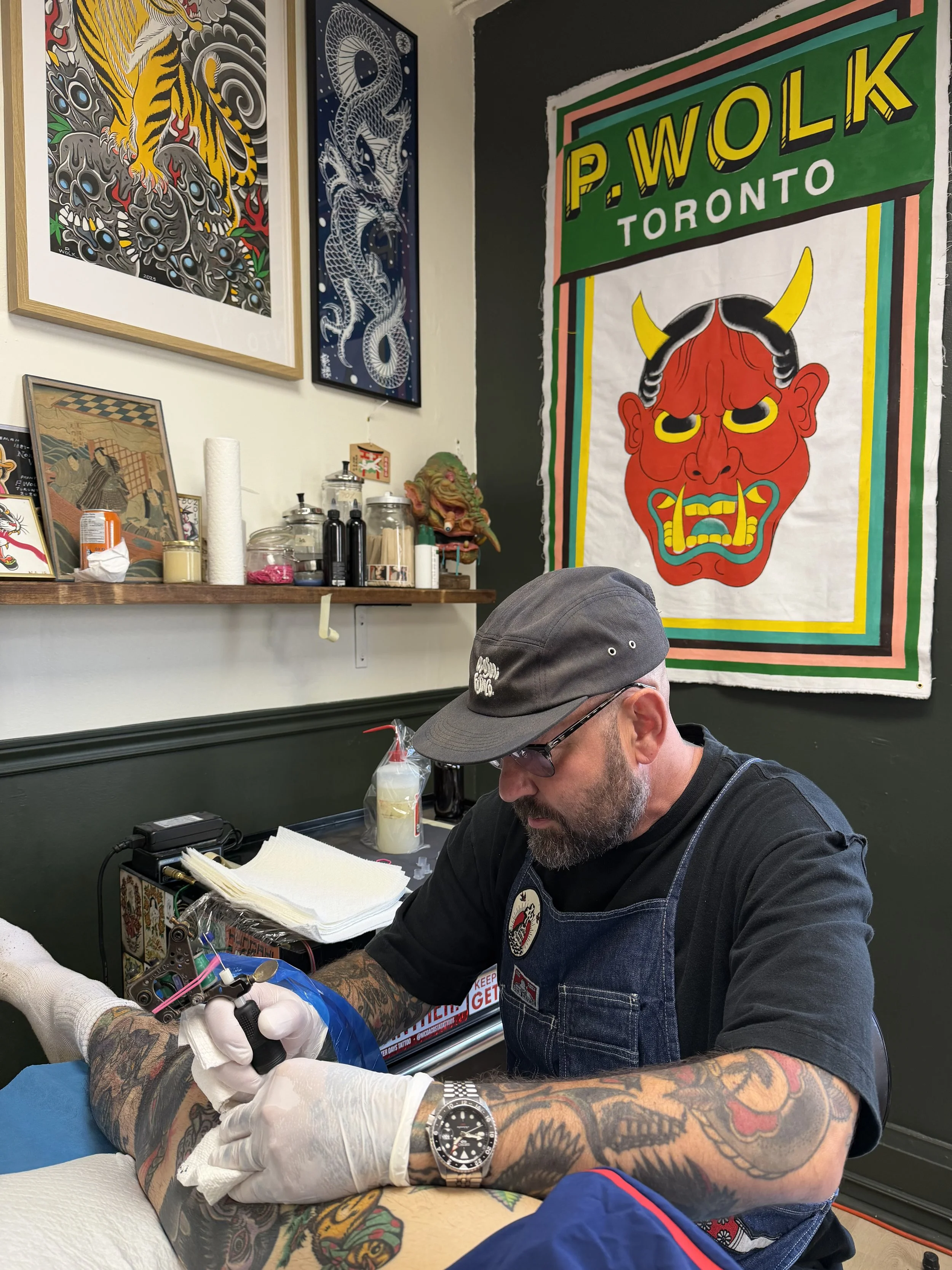 A man getting a tattoo on his arm at a tattoo parlor. He has tattoos on his arm and is wearing a gray cap, black t-shirt, and a denim vest. The tattoo artist's arm is visible, wearing gloves and working on the tattoo. The background features colorful artwork and posters on the wall, including a prominent red demon face with yellow horns, and other tattoo art and decorations.